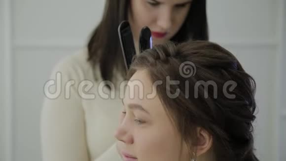 一位棕色头发的美女正在接受理发师用卷发熨斗治疗实时手持设备视频的预览图