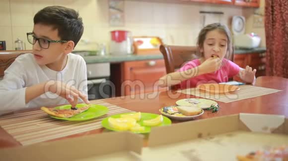 男孩和女孩双胞胎吃披萨和水果同时在餐桌旁的厨房里玩电话视频的预览图