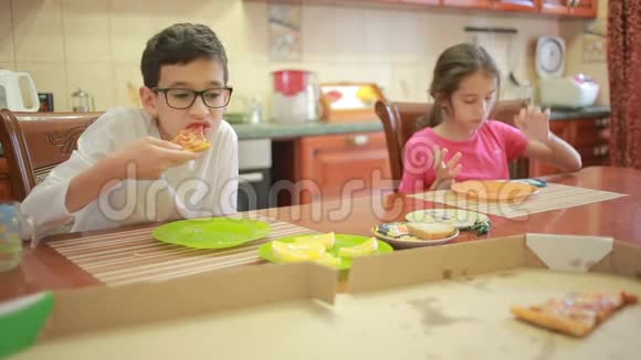 男孩和女孩双胞胎吃披萨和水果同时在餐桌旁的厨房里玩电话视频的预览图