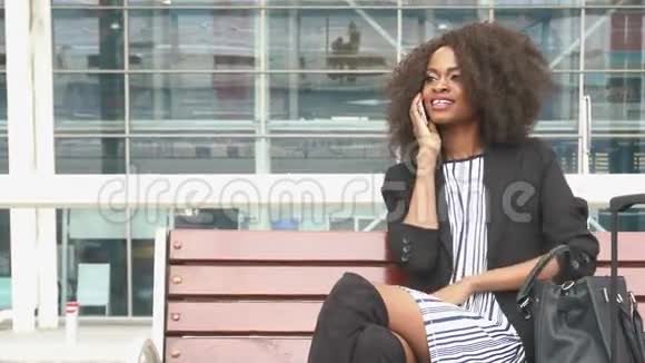 美丽微笑的非裔美国商务女性坐在机场的长凳上一边用手机说话视频的预览图