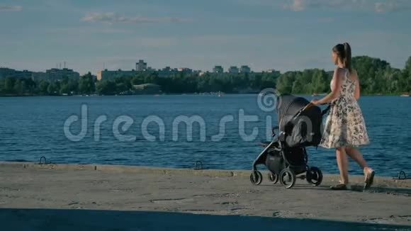 年轻漂亮的穿着衣服的妈妈带着婴儿车走在河边视频的预览图
