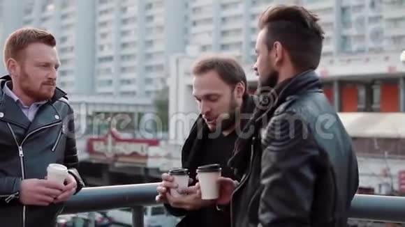 三个留着胡须的英俊青年在城市的一座桥栏杆附近交谈和喝咖啡慢节奏视频的预览图