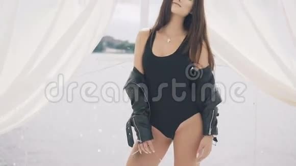 穿着黑色泳衣和夹克的漂亮女孩在海边的白色凉亭里摆姿势4K视频的预览图