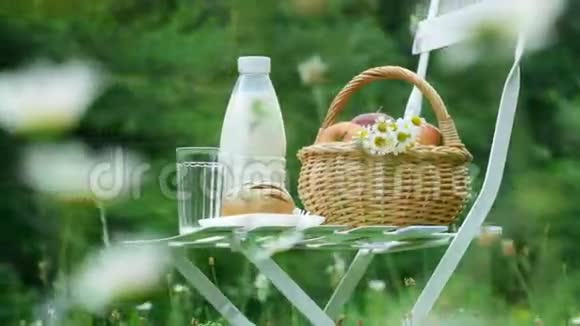 在洋甘菊草坪的中间白色的椅子上有一瓶牛奶一篮子苹果和面包视频的预览图