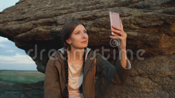 年轻的旅游女性背包客在日落时分在岩石上徒步旅行后在智能手机摄像头上拍摄风景视频的预览图