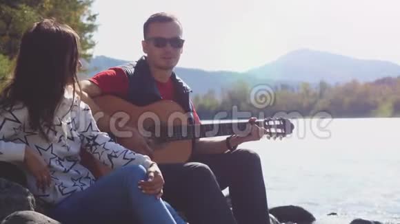 一对年轻夫妇在海边玩吉他唱歌在山河旁边的夏天3840x2160视频的预览图