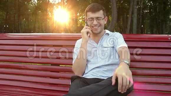一个戴太阳镜的年轻人坐在公园里用智能手机聊天视频的预览图