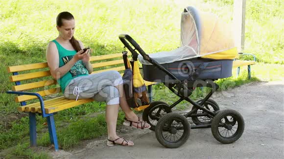 坐在公园里的母亲卷着婴儿车看着智能手机视频的预览图