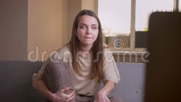 年轻迷人的白种人女性兴奋地坐在室内的沙发上看电视的特写镜头视频的预览图