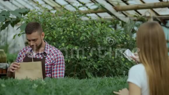 年轻微笑的卖花者在花园中心工作男人给顾客一个购物袋用信用卡付款视频的预览图