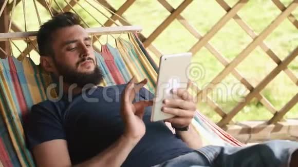 一个留胡子的年轻人用平板电脑躺在乡间别墅的吊床上视频的预览图