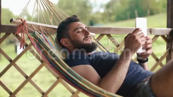 一个留胡子的年轻人用平板电脑躺在乡间别墅的吊床上视频的预览图