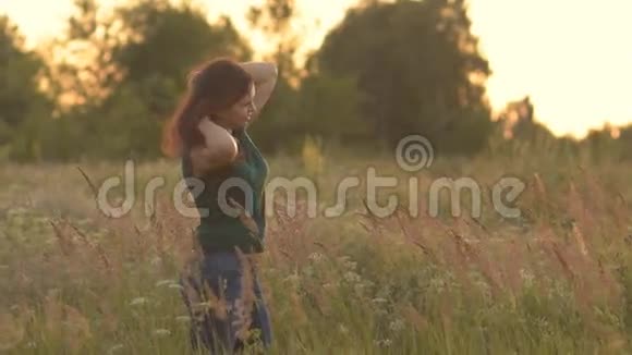 阳光下美丽的模特女孩穿着牛仔裤和绿色衬衫在田野上抚摸她的长发看着视频的预览图