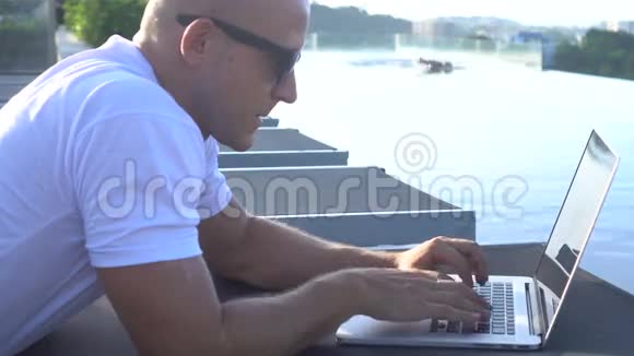 一个英俊的年轻人在笔记本电脑上工作躺在日光浴床上男人在无限泳池里游泳视频的预览图