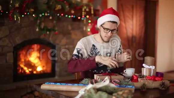 大胡子坐着在壁炉旁的新年礼物上系上蝴蝶结戴着圣诞帽的家伙视频的预览图