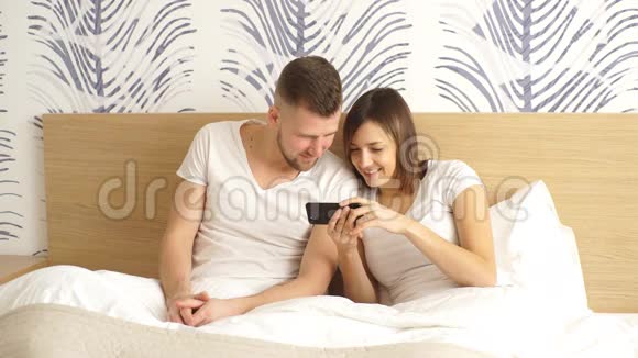 快乐的一对情侣在床上抱着智能手机在家里的卧室里视频的预览图