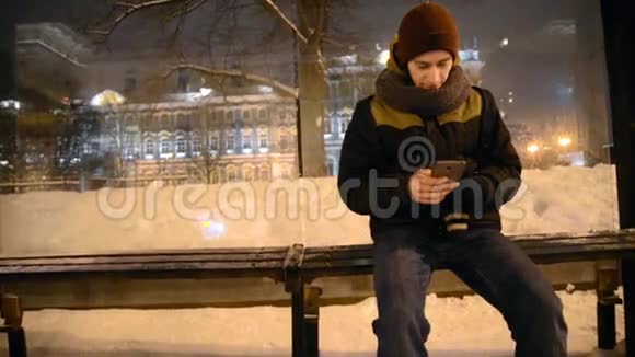 一个男人在一个灰色的阴天用智能手机坐在公共汽车站的特写镜头视频的预览图