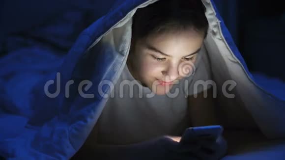 微笑少女躺在毯子下用手机的肖像视频的预览图