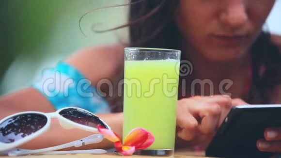 年轻有魅力的女人在海滩上用手机喝健康的果汁旁边是太阳镜和弗兰吉帕尼花视频的预览图