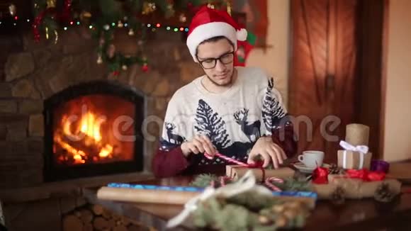 大胡子坐着在壁炉旁的礼物上打蝴蝶结戴着圣诞帽的人用纸包盒视频的预览图