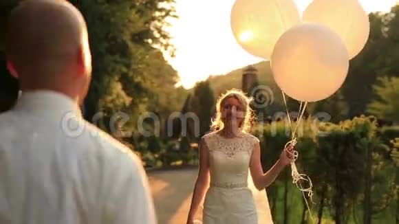 快乐的新娘和新郎一起散步在绿色的秋天公园里接吻手里拿着白色的气球金色夕阳视频的预览图