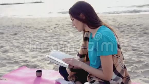 年轻女子在沙滩上看书喝热茶或热水瓶里的咖啡视频的预览图