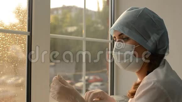 戴着口罩的女医生在窗户的背景上戴上橡胶手套视频的预览图