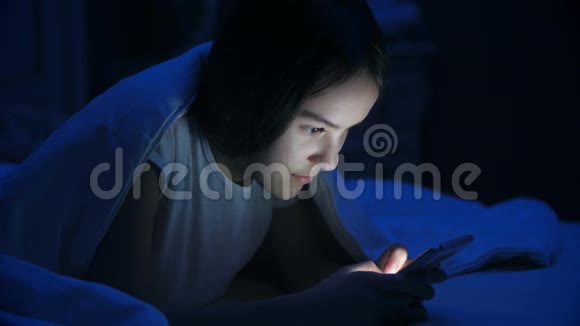 微笑少女躺在毯子下的肖像和手机打字信息视频的预览图