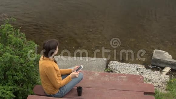 夏天一个年轻的女人坐在池塘边的码头上玩电话视频的预览图