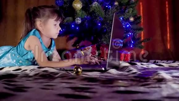 圣诞树下有笔记本电脑的女孩在新年里孩子在一棵树下手提电脑一个小女孩视频的预览图