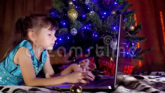 圣诞树下有笔记本电脑的女孩在新年里孩子在一棵树下手提电脑一个小女孩视频的预览图