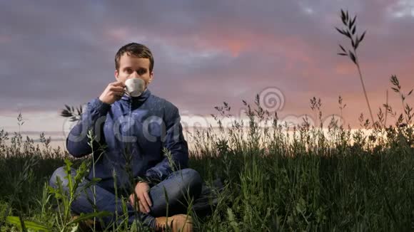 一个可爱的年轻人在田野日落时喝一杯热饮料背景上美丽的天空视频的预览图
