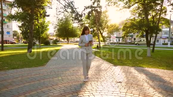 美丽的女商人走在公园的小巷里手里拿着平板电脑穿着轻便商务服的女孩去上班视频的预览图