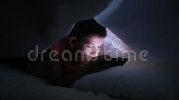 在床上玩手机或智能手机的男孩躲在毯子下面视频的预览图