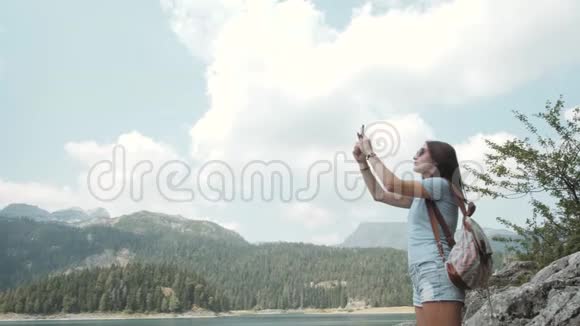 年轻女子用智能手机在山湖前拍照美丽的白种人女孩在一个女孩子中度过的时光视频的预览图