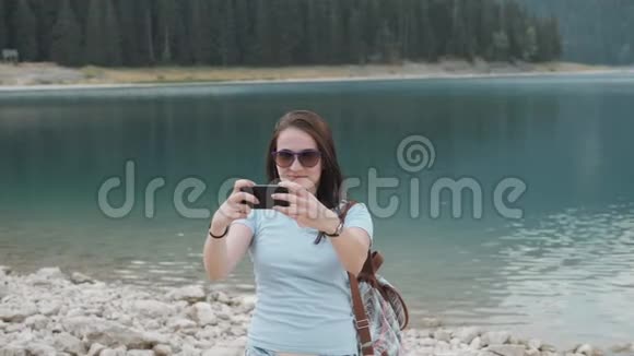 年轻女子在山湖前用智能手机自拍美丽的白种人女孩在一个女孩子中度过的时光视频的预览图