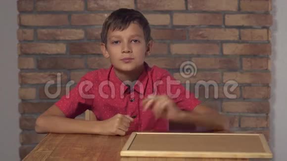 坐在书桌前的孩子拿着一张挂着字母的挂图在背景红砖墙上打电话给我视频的预览图