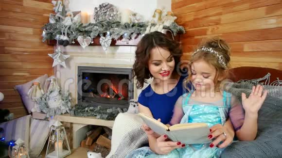 母亲和女儿读一本书一家人坐在壁炉和圣诞树旁看圣诞故事新视频的预览图