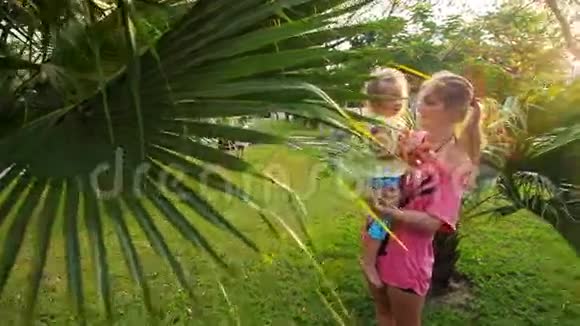 妈妈抱着胳膊女儿看公园里的棕榈树视频的预览图