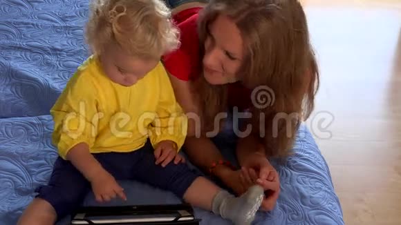 金发碧眼的婴儿和黑发女人妈妈在蓝色的床上用药片视频的预览图