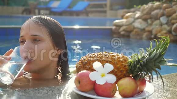 年轻女子一边喝水一边在池中晒太阳盘子里放着水果和梅花动作缓慢视频的预览图