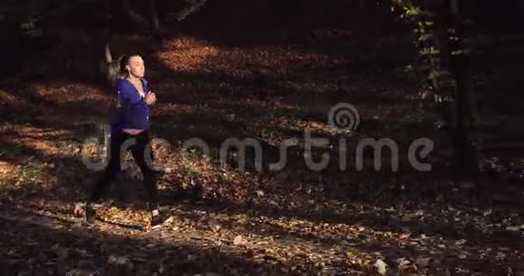 公园里的女跑步者穿着蓝色夹克和其他健身服的漂亮女人跑步时听音乐视频的预览图