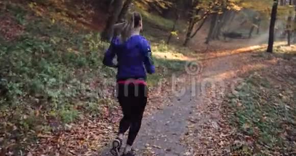 公园里的女跑步者穿着蓝色夹克和其他健身服的漂亮女人跑步时听音乐视频的预览图