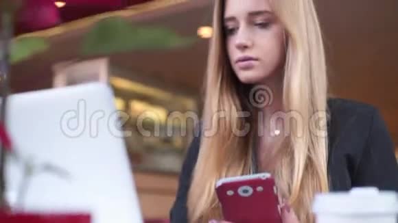 穿着黑色夹克的漂亮金发女郎正坐在咖啡馆里她正在用她的智能手机和笔记本视频的预览图