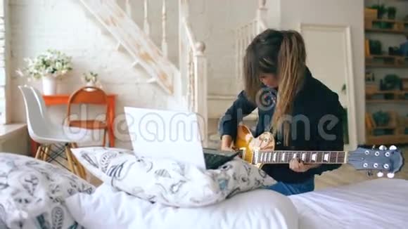 有魅力的年轻女孩在家里的卧室里坐在床上学习用笔记本弹电吉他视频的预览图