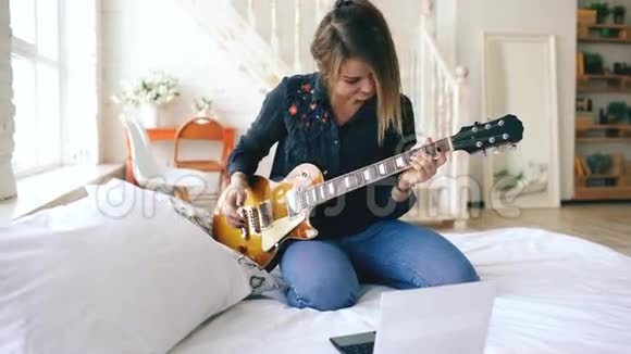 有魅力的年轻女孩在家里的卧室里坐在床上学习用笔记本弹电吉他视频的预览图