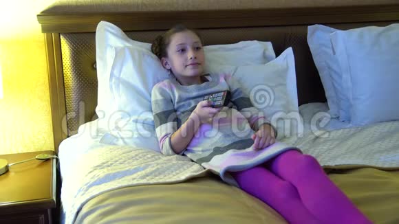 漂亮的小女孩坐在旅馆的床上看电视手里拿着遥控器的女孩视频的预览图