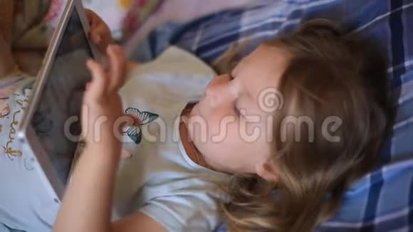 小白种人两岁的女孩躺在格子枕头上在平板电脑上看动画片现代技术视频的预览图