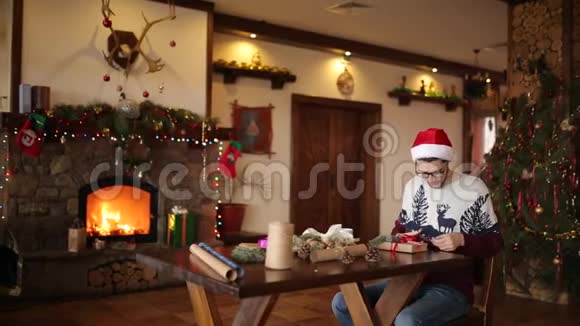 大胡子坐在壁炉旁的礼物上打蝴蝶结戴着纸包圣诞帽装饰着视频的预览图