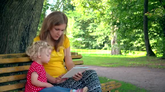 母亲带着蹒跚学步的女儿女孩用平板电脑坐在公园的长凳上视频的预览图